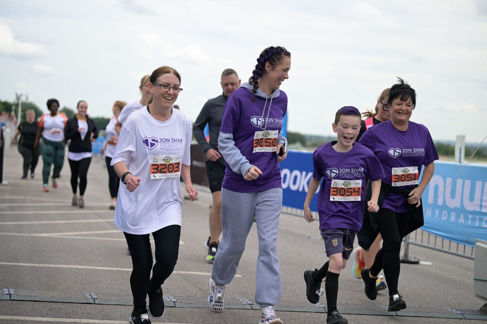 A group of individuals wearing running gear participates in a race event. Some are wearing purple shirts with race bibs, while others wear white shirts. They appear to be walking or jogging along a paved path, smiling and enjoying the activity. The sky is overcast.