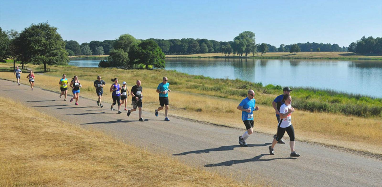A group of people running on a paved path along a scenic lake. The runners are dressed in various athletic wear, and the path is bordered by grassy areas and trees. The sky is clear and blue, suggesting a bright, sunny day.