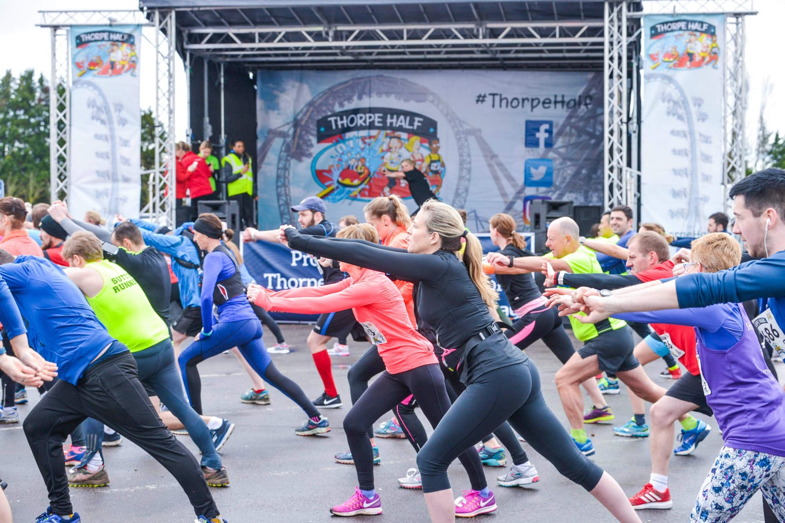 A group of runners in colorful athletic wear performing warm-up exercises in front of a stage with the banner "Thorpe Half" during an organized race event. Banners and some onlookers are visible in the background.