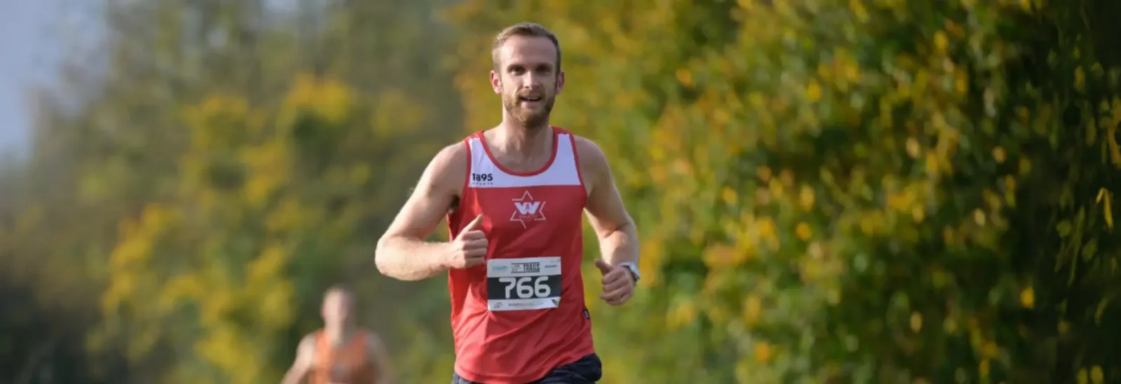 A man in a red running vest with race number 766 is running outdoors on a sunny day, giving a thumbs-up gesture, with blurred green trees in the background.