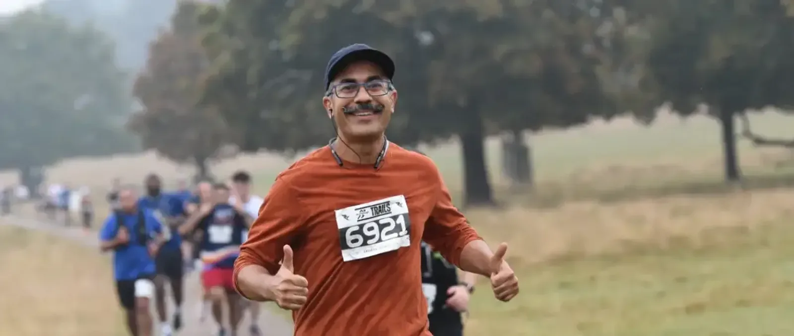 A smiling man wearing glasses, a cap, and an orange shirt with race bib 6921 runs outdoors, giving two thumbs up. Other runners and trees are visible in the background.
