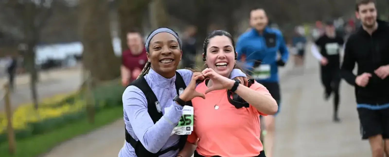 Two women are smiling and forming a heart shape with their hands while running in a race, with other runners and trees visible in the blurred background.