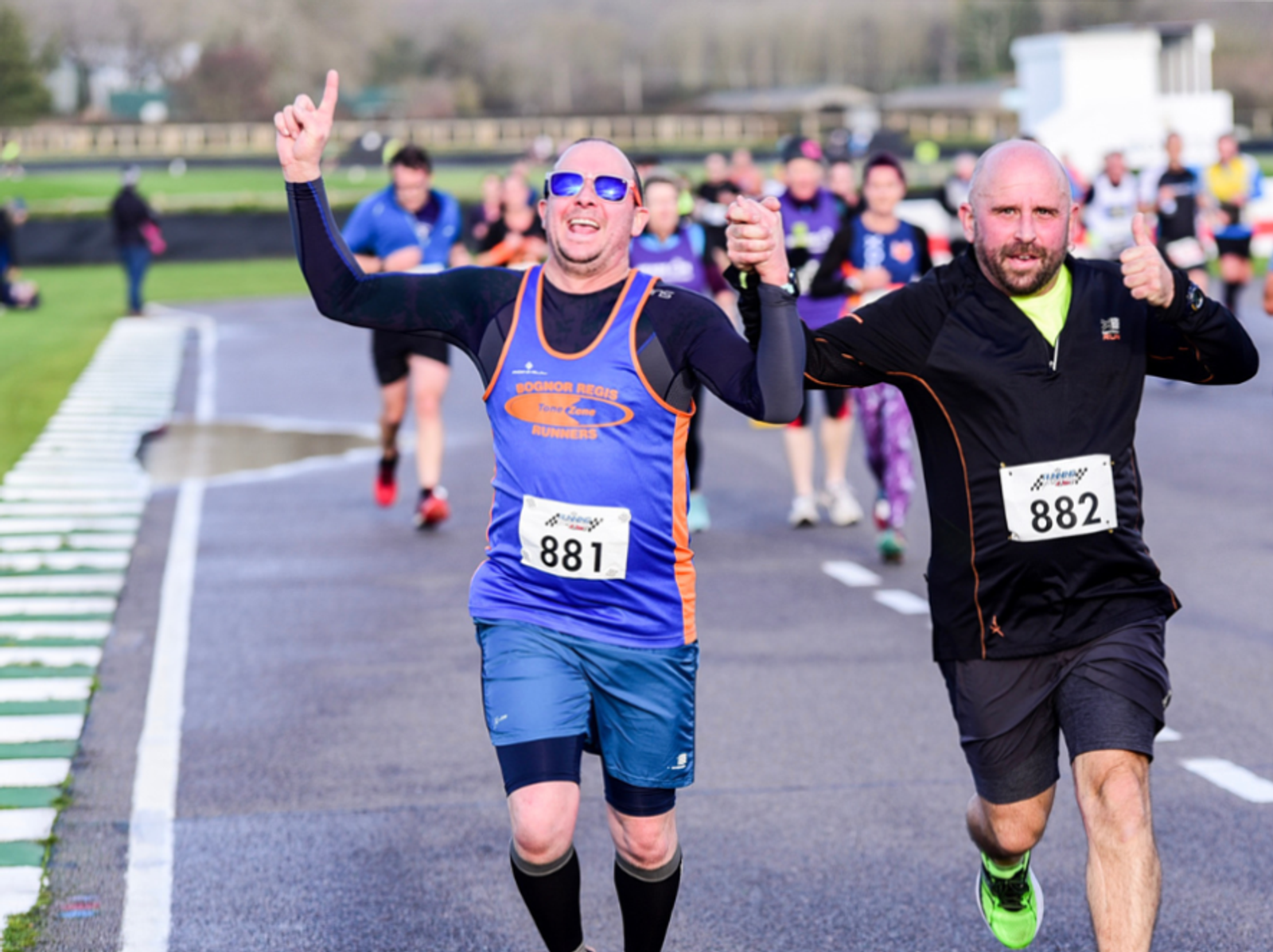 Two men, wearing race bibs numbered 881 and 882, cheer as they run on a road, surrounded by other runners. They hold hands and display expressions of joy. The event takes place on a clear day with green fields in the background.