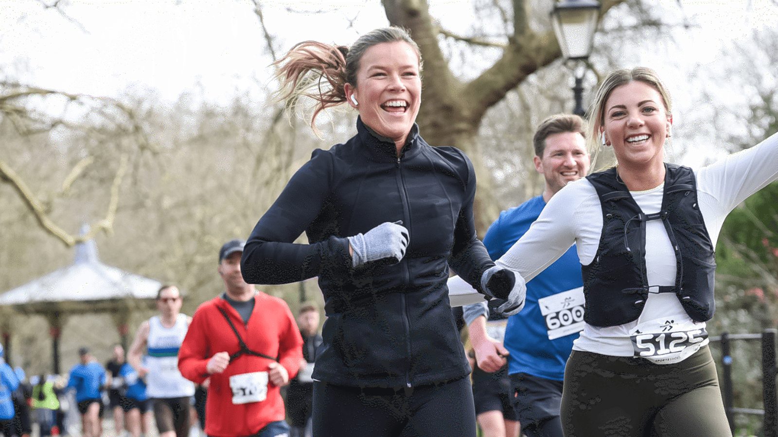 Two women wearing athletic gear run side by side, smiling energetically during a race in a park. In the background, other runners are visible against a backdrop of leafless trees and a gazebo.