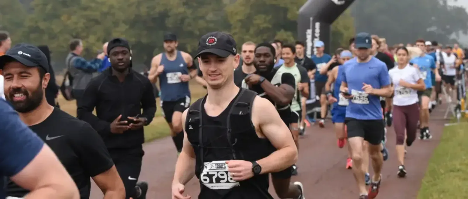 A group of runners, both men and women, are participating in a race on a paved path outdoors, surrounded by trees. Most wear athletic gear and race bibs, with some smiling as they run.