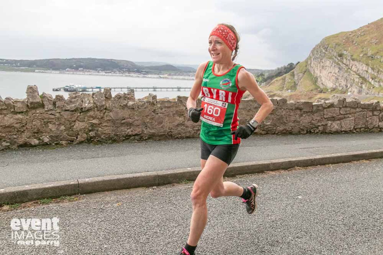 A runner in a red and green outfit, bib number 160, smiles during a road race near a coastal area. The background features hills and the sea, while the roadway is partially bordered by a stone wall. The logo "event IMAGES" is visible in the bottom left corner.
