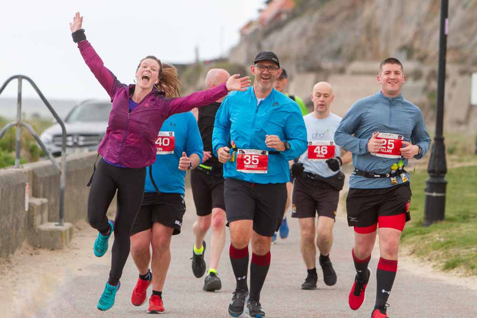 A group of runners participating in a race along a seaside path. The runner in the foreground, wearing a purple jacket, is leaping joyfully with arms raised. Others around her, dressed in blue and grey athletic gear, are running with focused expressions.