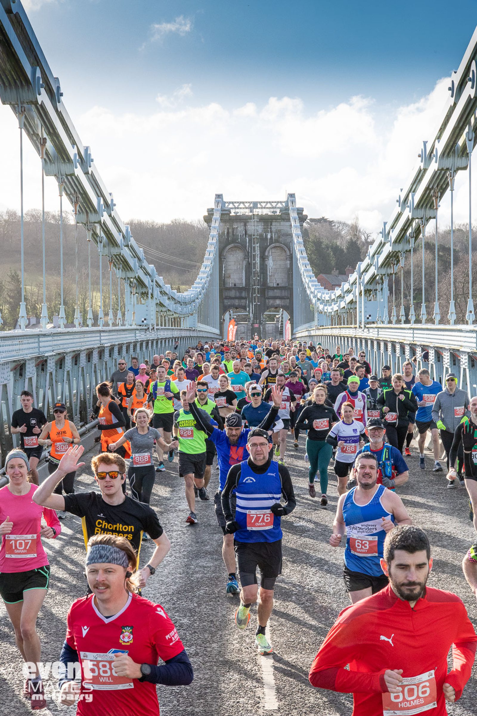 A large group of runners participate in a race on a suspension bridge. The crowd displays a mix of colorful athletic attire and numbered bibs. In the background, the structure of the bridge and a partly cloudy sky are visible. Some runners wave or smile at the camera.