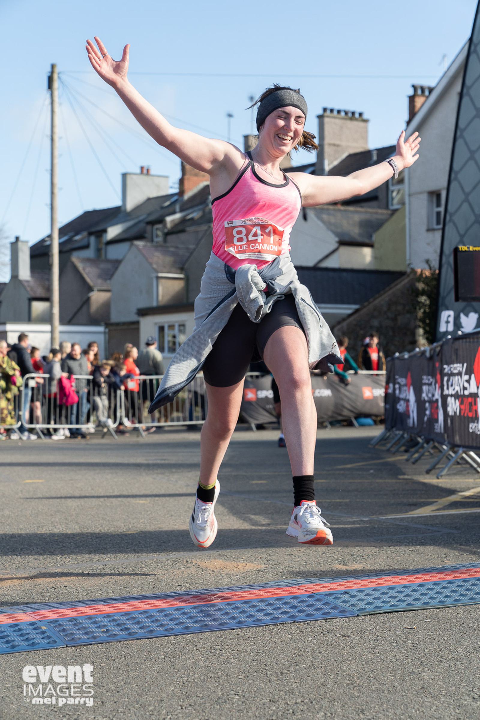 A woman in athletic wear joyfully leaps across a finish line during a race on a sunny day. She has her arms outstretched and is smiling. There are buildings and a crowd cheering in the background. The ground is marked with an orange and blue stripe.