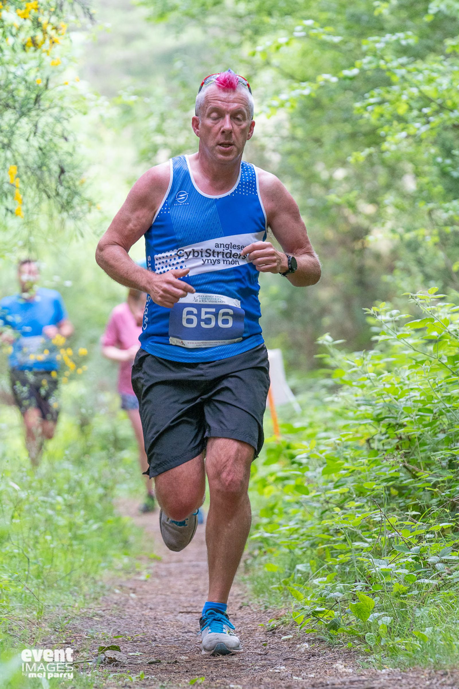 A middle-aged man with a pink mohawk haircut runs on a forest trail. He's wearing a blue running tank top with white accents, black shorts, and sports a race bib with the number 656. Trees and greenery surround the trail. Another runner is visible in the background.