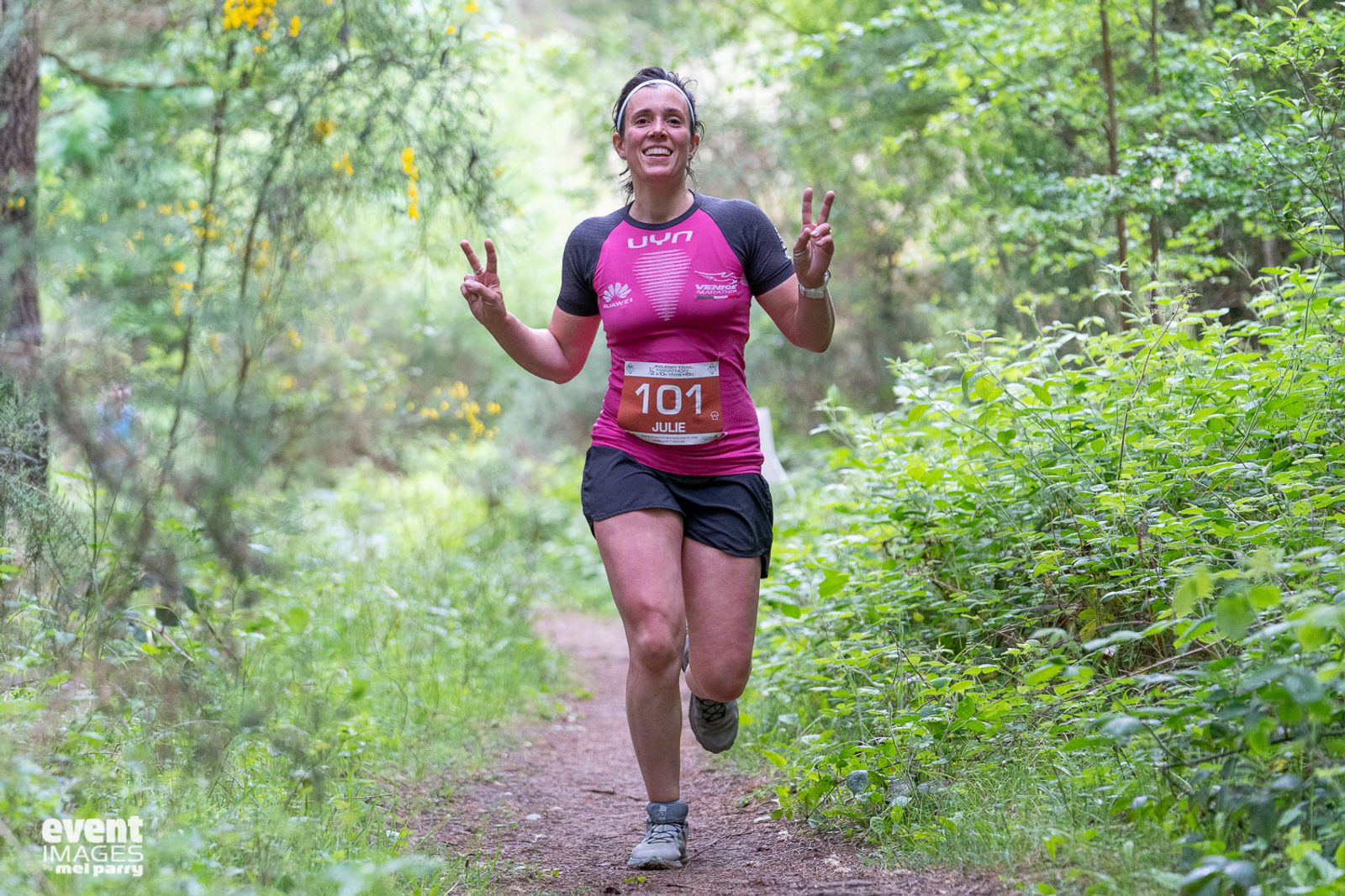 A woman wearing a pink and black running outfit, bib number 101, runs along a forest trail, smiling and holding up two peace signs with both hands. The path is surrounded by lush green foliage. The weather appears warm and sunny. Text in the bottom left corner reads "event IMAGES".