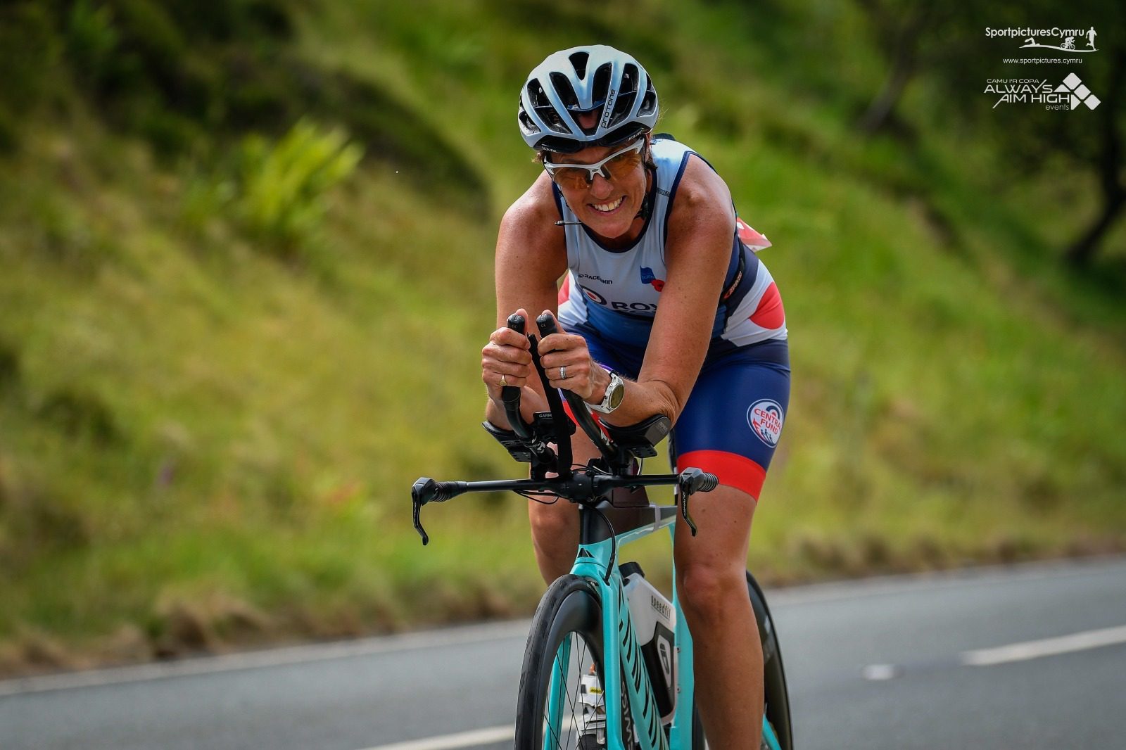 A cyclist wearing a blue and red triathlon suit and helmet rides a teal bicycle on a paved road. The road is flanked by green foliage, and the athlete seems focused on maintaining speed. Logos from various sponsors are visible on the image.