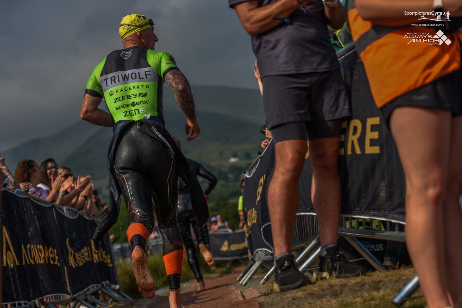 A triathlete in a green and black wetsuit runs along a race course, past cheering spectators and race officials. Mountains and a cloudy sky are visible in the background. The athlete's back is to the camera, highlighting various sponsor logos on their outfit.