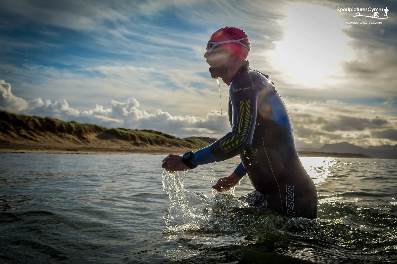 A person in a wetsuit and red swim cap emerges from a body of water, with water cascading down their arms. They are wearing goggles and a watch. The background features a sandy shoreline, grassy dunes, and a dramatic sky with the sun partially covered by clouds.