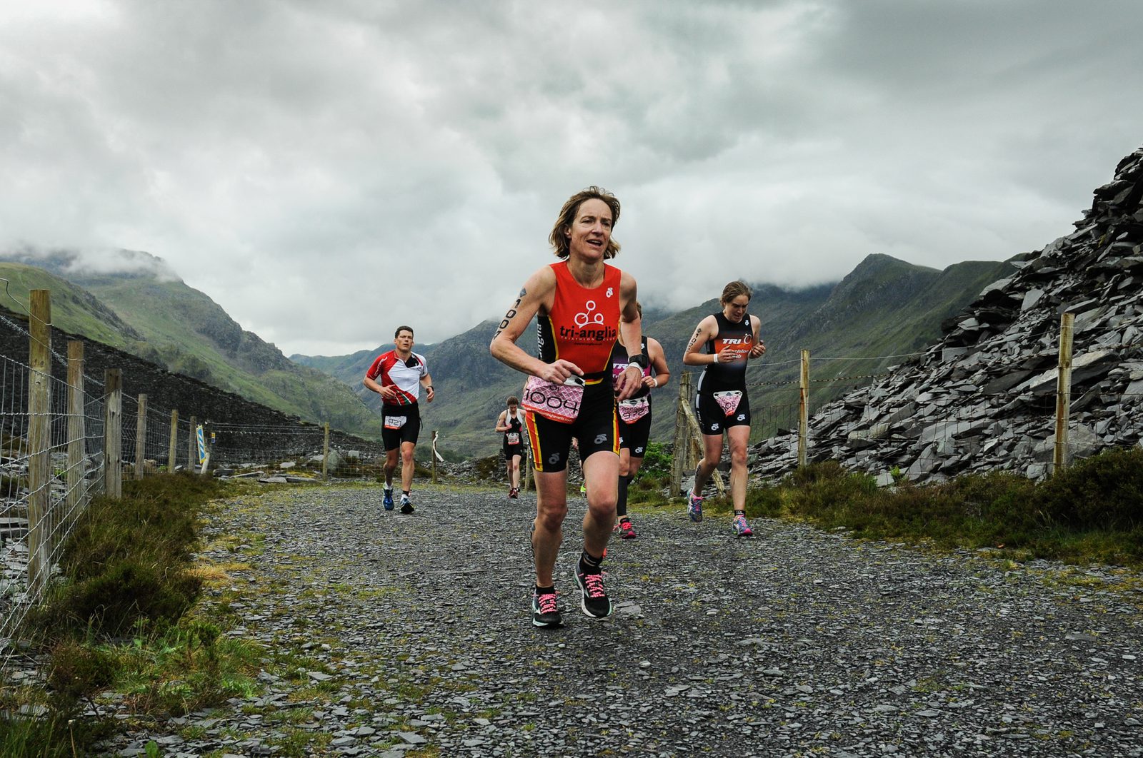 A group of athletes in triathlon gear run on a gravel path surrounded by hills and a cloudy sky. A barbed wire fence is on the left, and a rocky slope is on the right. The lead runner, wearing a red top, appears to be exerting significant effort.