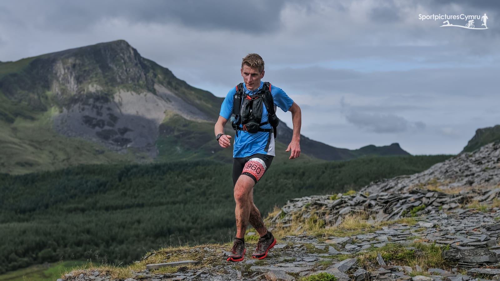 A man in a blue shirt, bib number 6936, runs on a rocky trail with a scenic mountainous backdrop. He appears focused and determined, competing in an outdoor trail running event. The landscape includes green forested areas and a cloudy sky.