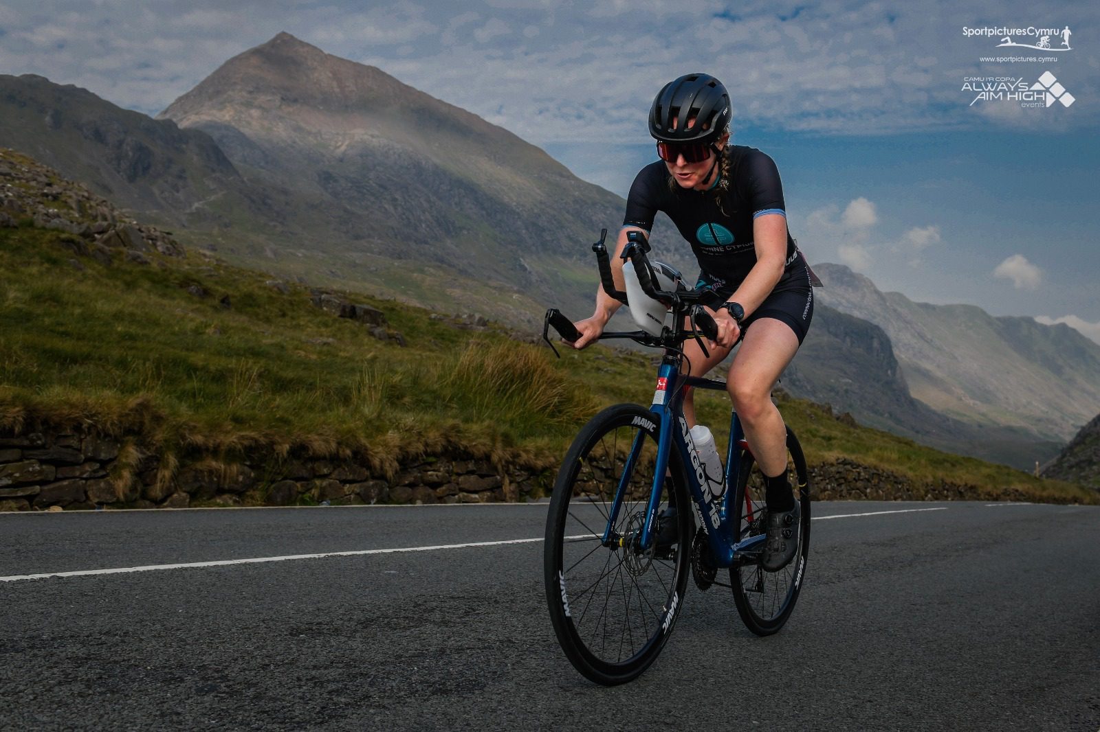 A cyclist in black and blue gear rides a road bike up a mountainous road. The backdrop features rugged mountains under a partly cloudy sky. The cyclist wears a helmet and sunglasses, focused on the climb. Logos are visible in the top right corner of the image.