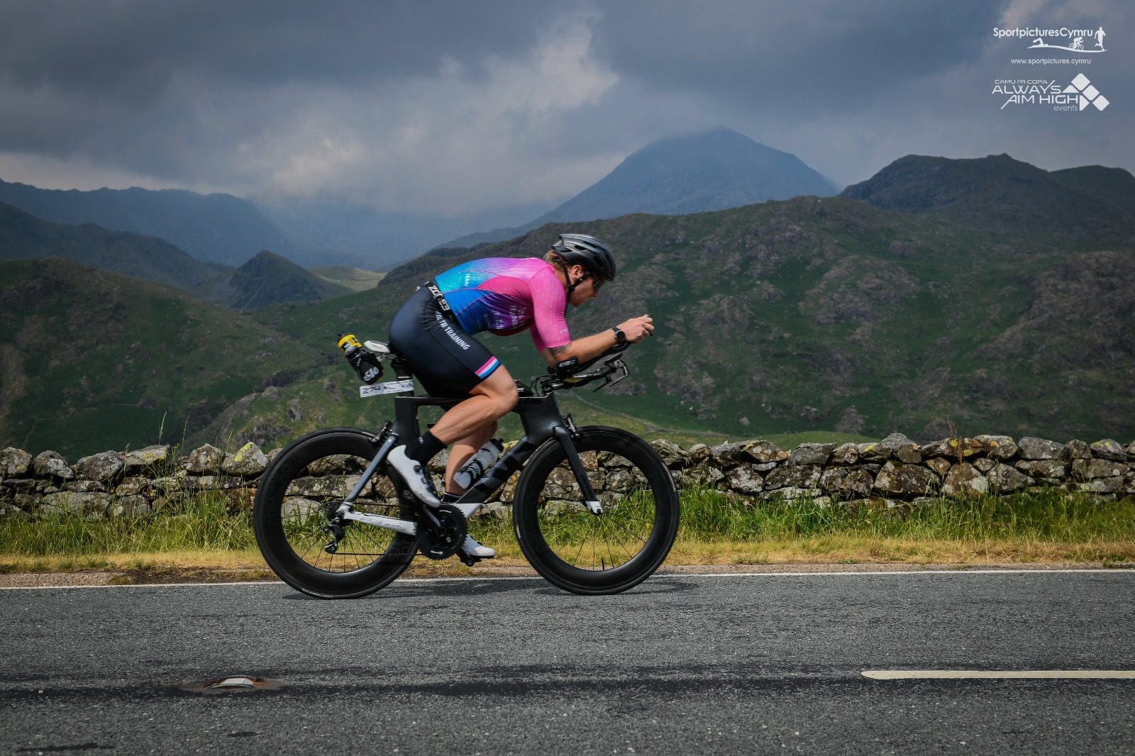 A cyclist wearing a blue and pink jersey rides a road bike along a mountainous landscape. They are in a crouched position on the bike, with a stone wall and green hills in the background under a cloudy sky.