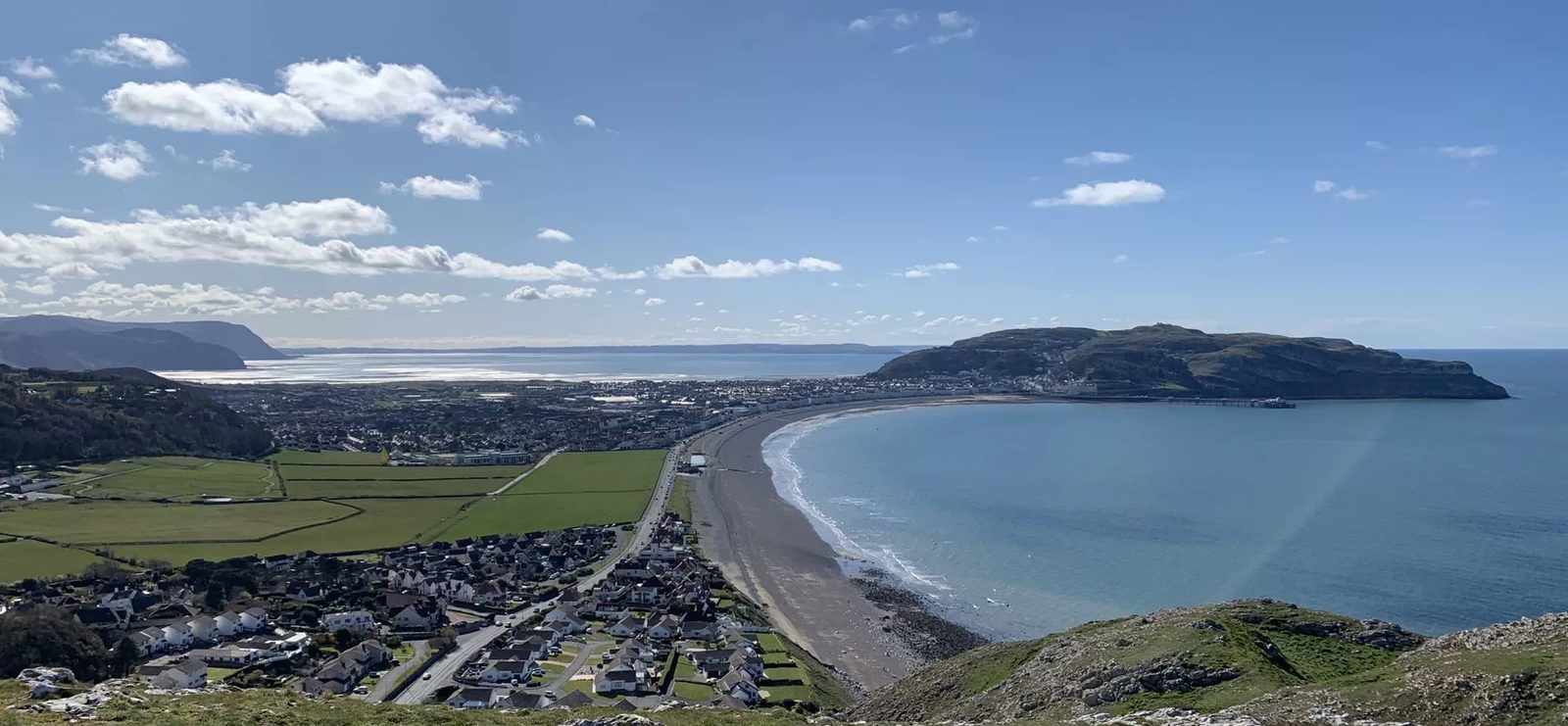 A panoramic view of a coastal town featuring a curved beach, calm blue waters, and a stretch of residential houses and green fields. Hills and mountains can be seen in the distance under a clear, sunny sky with scattered white clouds.