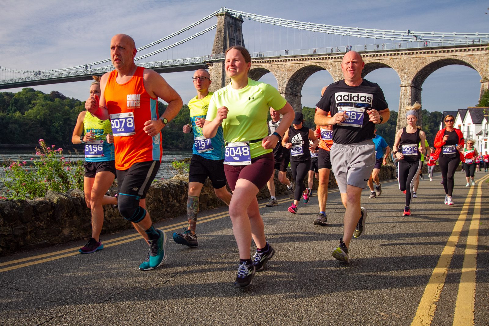 A group of runners participates in a marathon on a sunny day, with a historic suspension bridge in the background. The runners, wearing various athletic attire and numbered bibs, appear focused and determined as they race along a waterfront road.