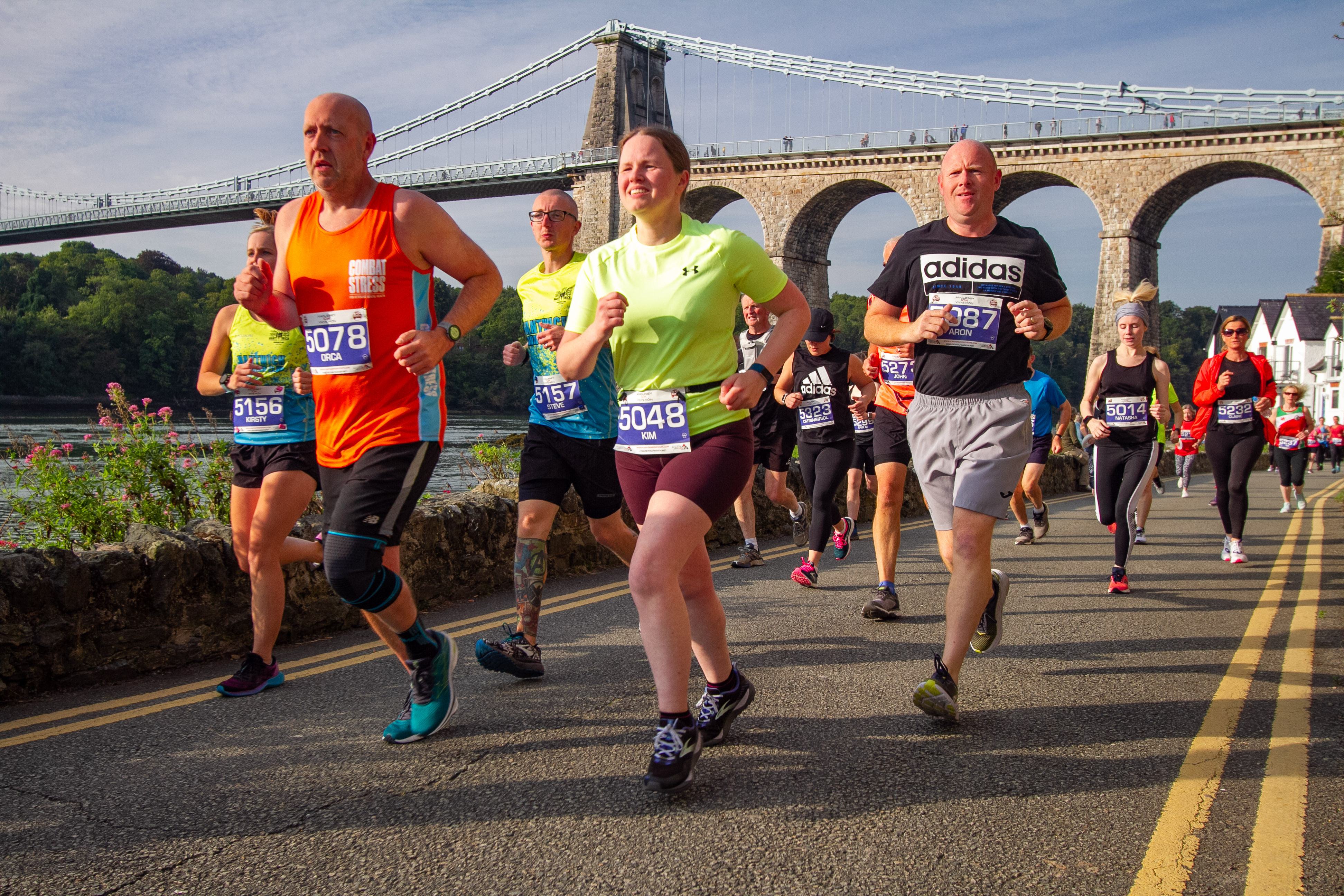 A group of runners participates in a marathon on a sunny day, with a historic suspension bridge in the background. The runners, wearing various athletic attire and numbered bibs, appear focused and determined as they race along a waterfront road.