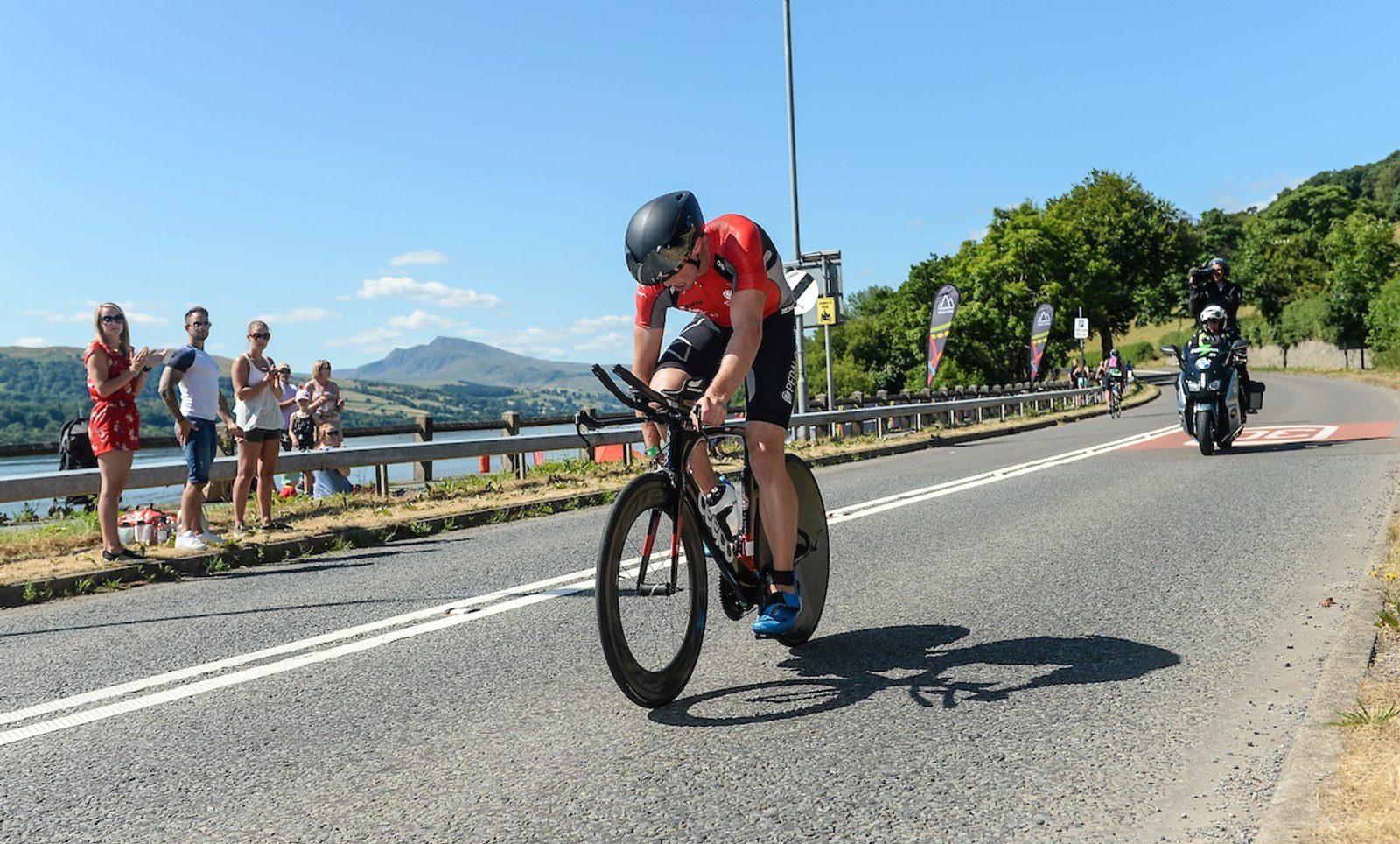A cyclist wearing a red jersey and black helmet speeds down a road on a time trial bike. Spectators stand on the side cheering, and a motorcyclist follows behind. The scene takes place on a sunny day with a scenic background of hills and trees.