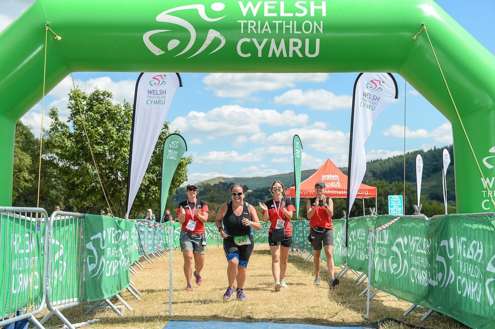 A triathlete runs towards the finish line of the Welsh Triathlon Cymru under a green inflatable arch. She is smiling and giving a thumbs-up sign, while two volunteers stand on either side, clapping and cheering. Flags and banners with the event logo are visible.