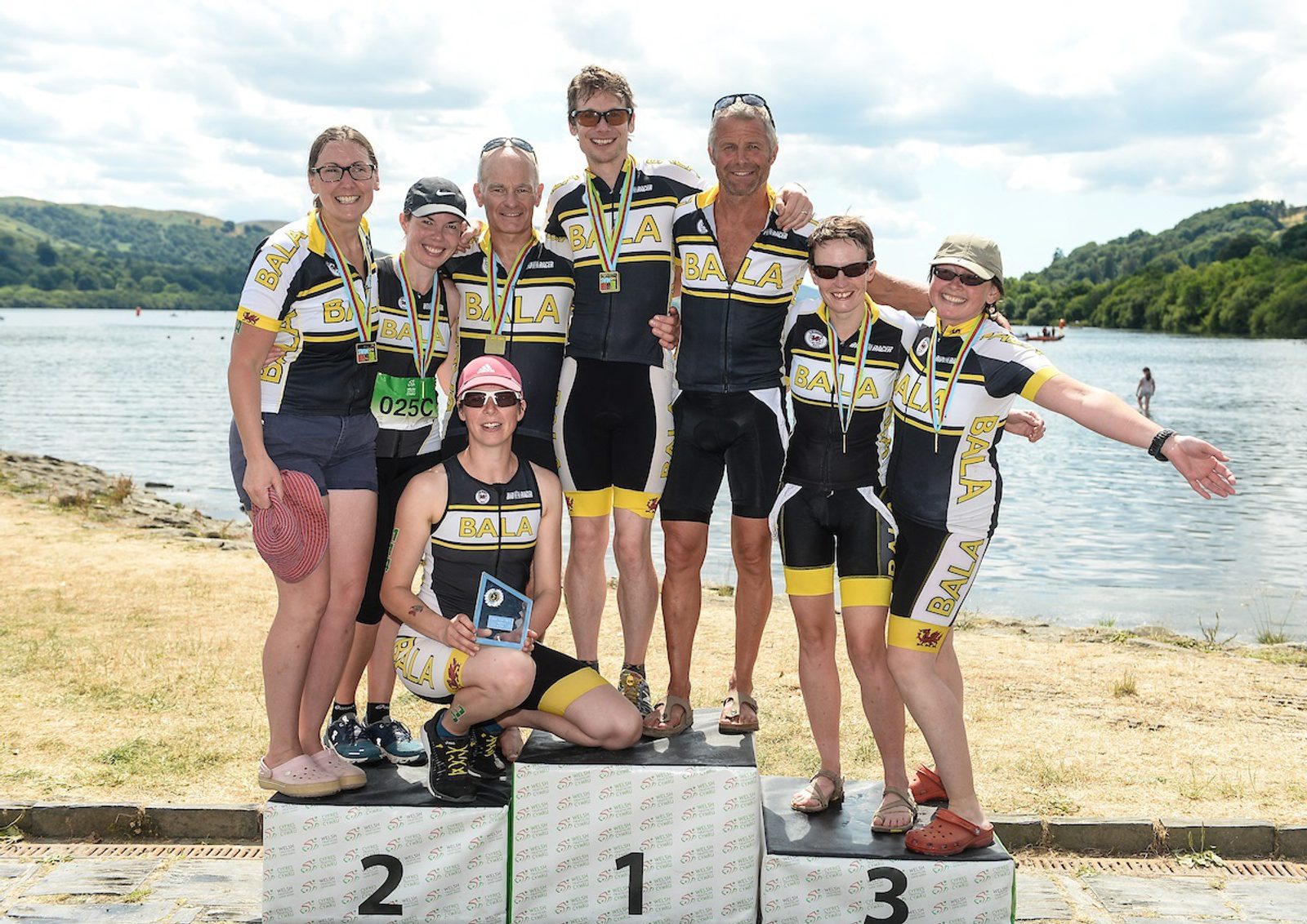 A group of eight smiling triathletes in matching yellow and black Bala uniforms stand and sit on podiums labeled 1, 2, and 3 by a lakeside. They are wearing medals, and one holds a trophy. The background includes a scenic view of hills and water under a partly cloudy sky.