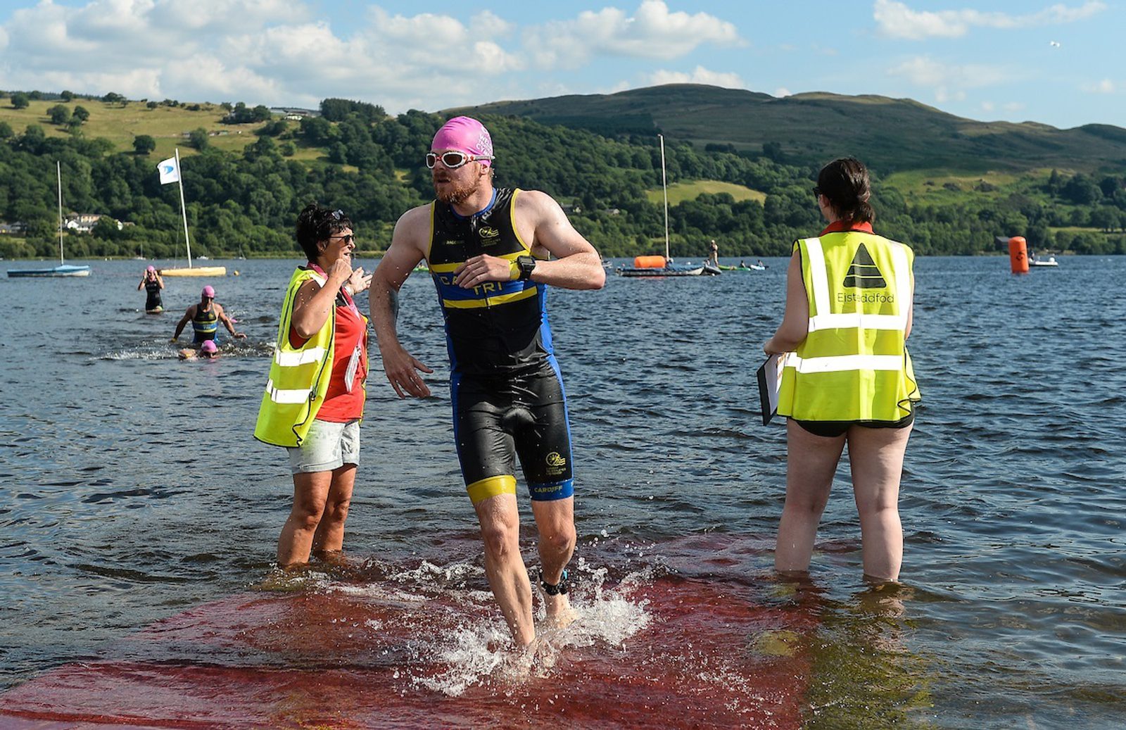A male triathlete in a black and blue wetsuit and pink swim cap runs out of the water onto a red mat during a triathlon, while two volunteers in yellow vests assist and monitor him. Hills and sailboats are visible in the background under a partly cloudy sky.