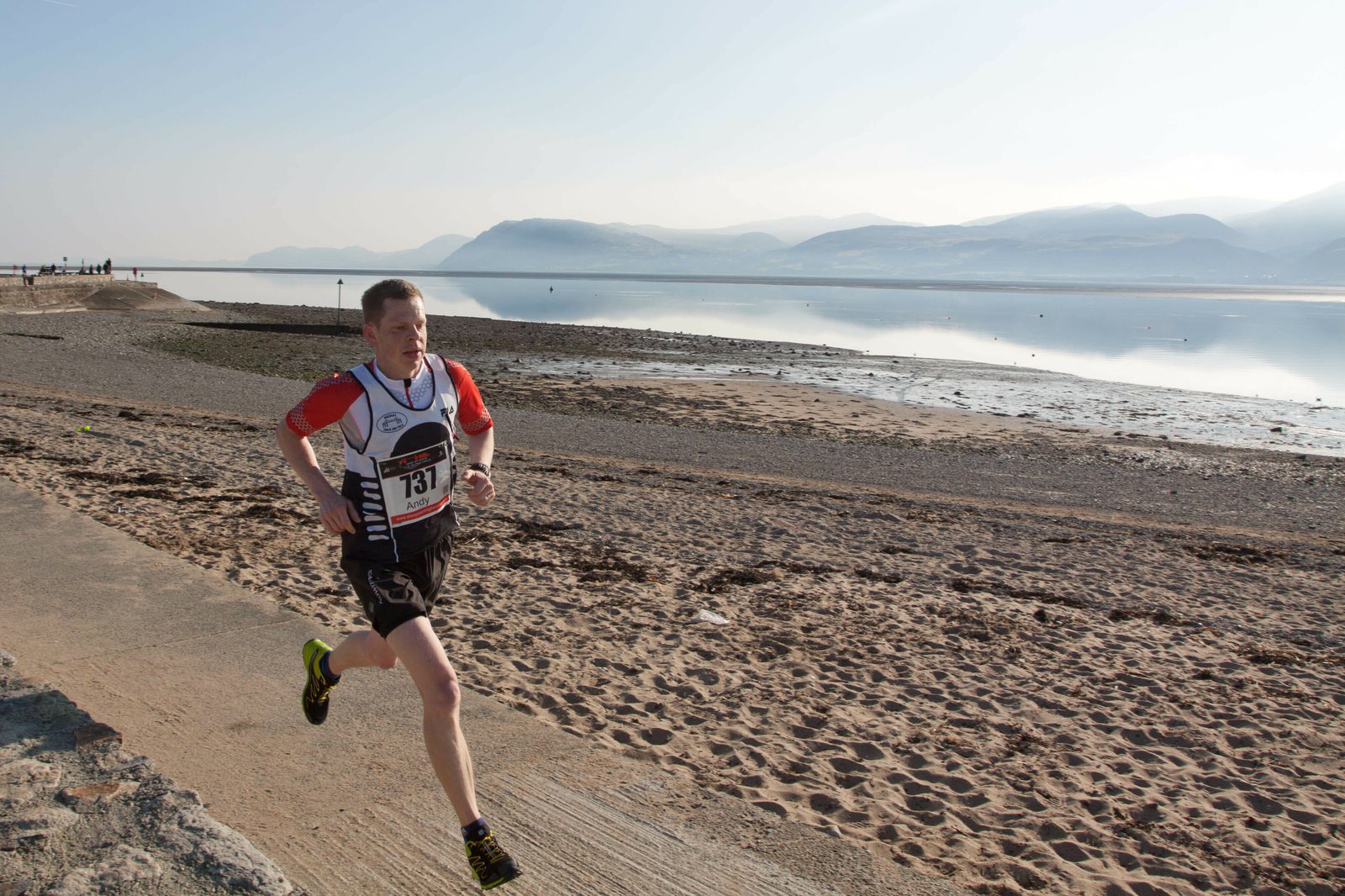 A person in a race bib with the number 731 is running along a sandy beach with a calm body of water and distant mountains in the background. They are wearing a red and white top and black shorts. The sky is clear and sunlit.