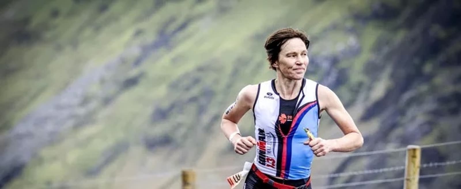 A person in a blue and white athletic outfit is running on a trail with a mountainous landscape in the background. The runner appears focused and determined, with the shirt featuring a distinctive design and logo. The scene captures an outdoor endurance event.