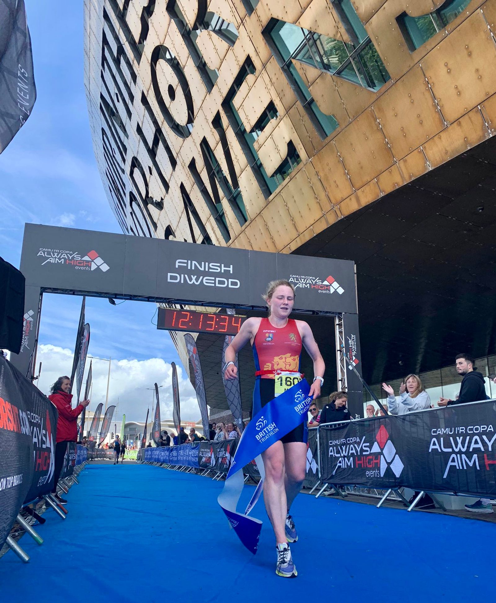 A runner wearing a red and yellow triathlon suit crosses the finish line triumphantly, holding a blue race banner. Spectators cheer on both sides, and the impressive architecture of the Wales Millennium Centre looms in the background. The displayed time reads 1:21:33.