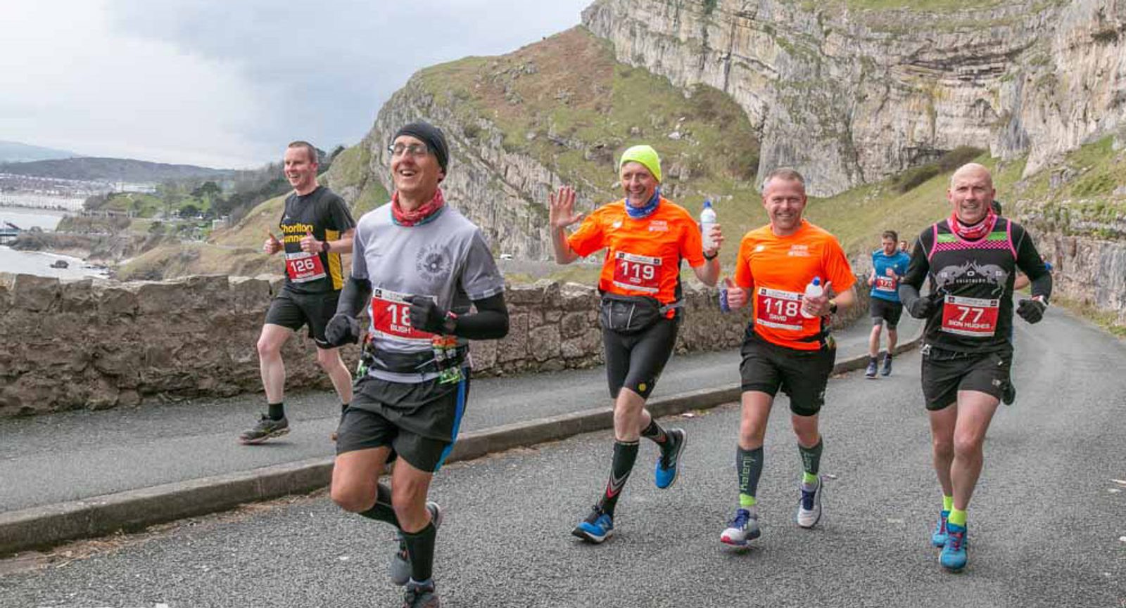 A group of six runners, dressed in bright athletic gear and numbered bibs, are running along a coastal road with rocky cliffs and the ocean in the background. They appear to be participating in a race, and most of them are smiling or waving.