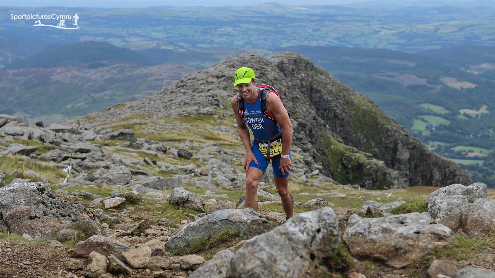 A person is climbing a rocky mountain trail, wearing athletic gear and a bright green cap. The backdrop features expansive, lush green hills and a cloudy sky. A logo in the top left corner reads "SportpicturesCymru.