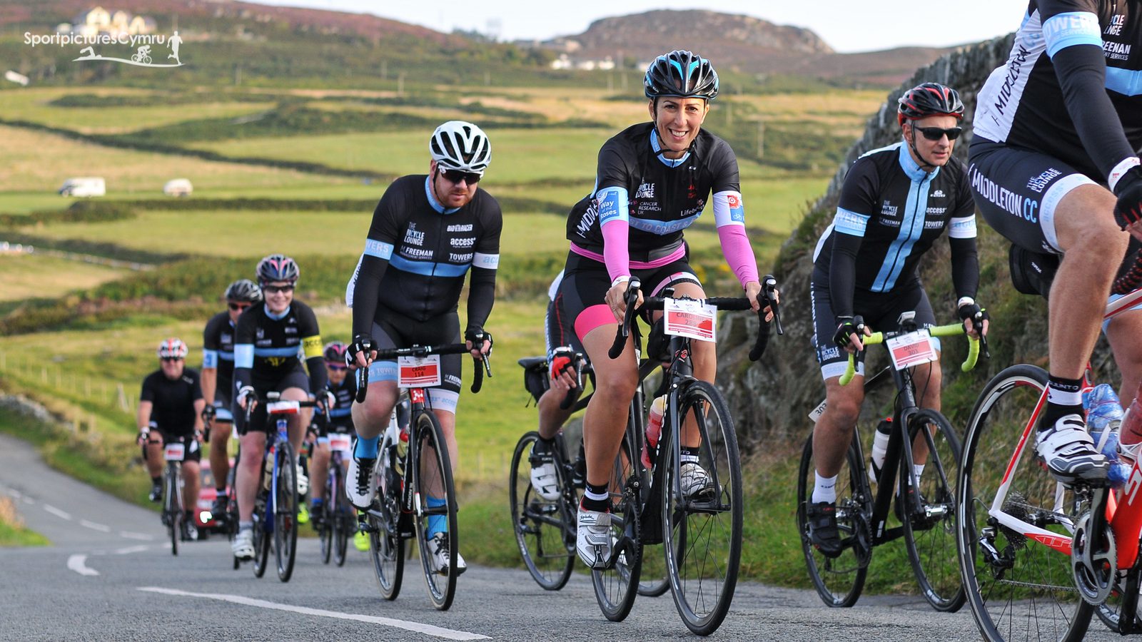 A group of cyclists ride up a winding road through a picturesque countryside featuring green fields and rolling hills. The cyclists are dressed in various biking gear, including helmets and jerseys. A smiling cyclist in the front wears a black and pink outfit. The sky is clear.