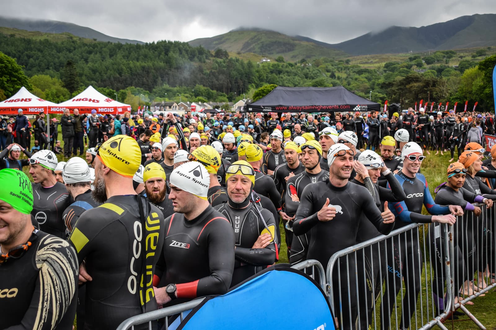 A large group of triathletes dressed in wetsuits and swim caps stand behind a metal barrier, preparing for a triathlon swim segment. The background features lush green hills and overcast skies. Tents and more racers are visible in the distance.