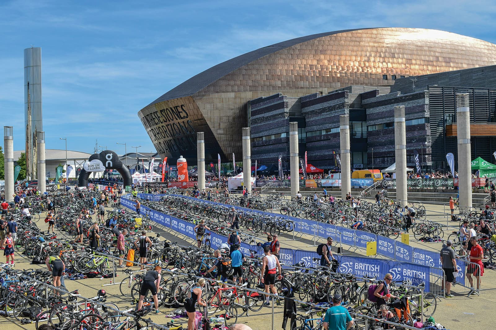Hundreds of bicycles and athletes are gathered in a busy outdoor transition area for the British Triathlon event. People are seen preparing their gear, with a modern, architecturally distinctive copper-colored building in the background under a clear blue sky.