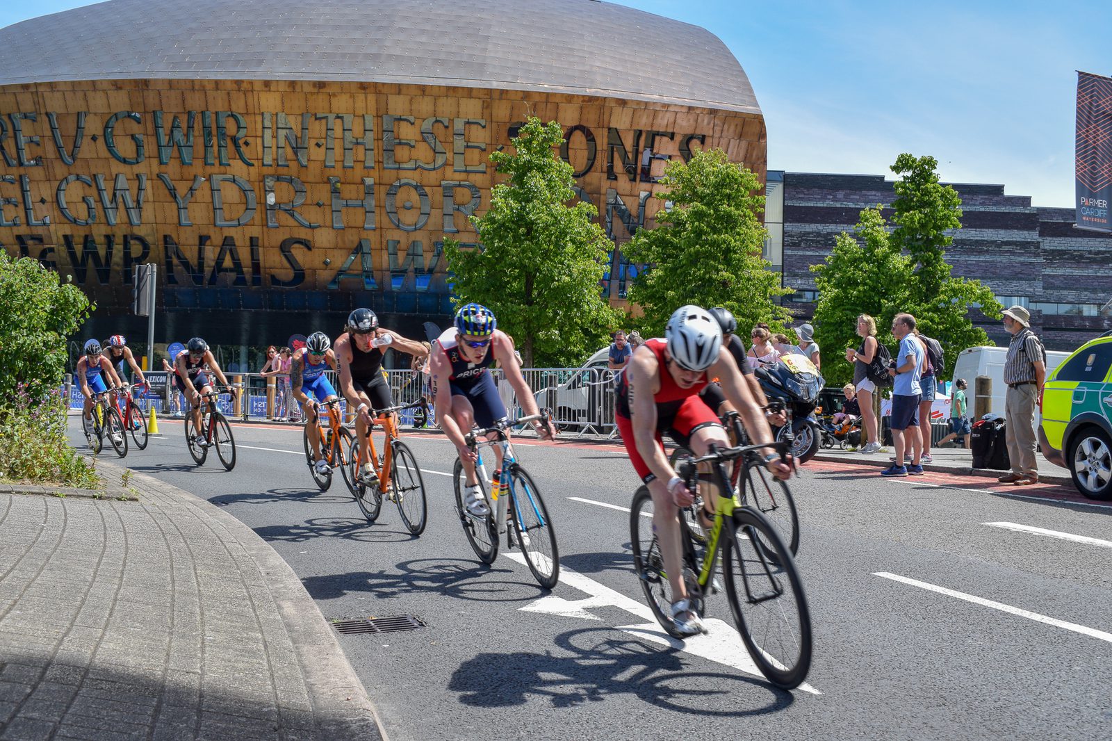 A group of cyclists participating in a race pass by a distinctive modern building with large, golden letters on its facade. Spectators watch from the roadside, while a medic and their motorcycle are on standby. Trees and a partly cloudy sky can be seen in the background.