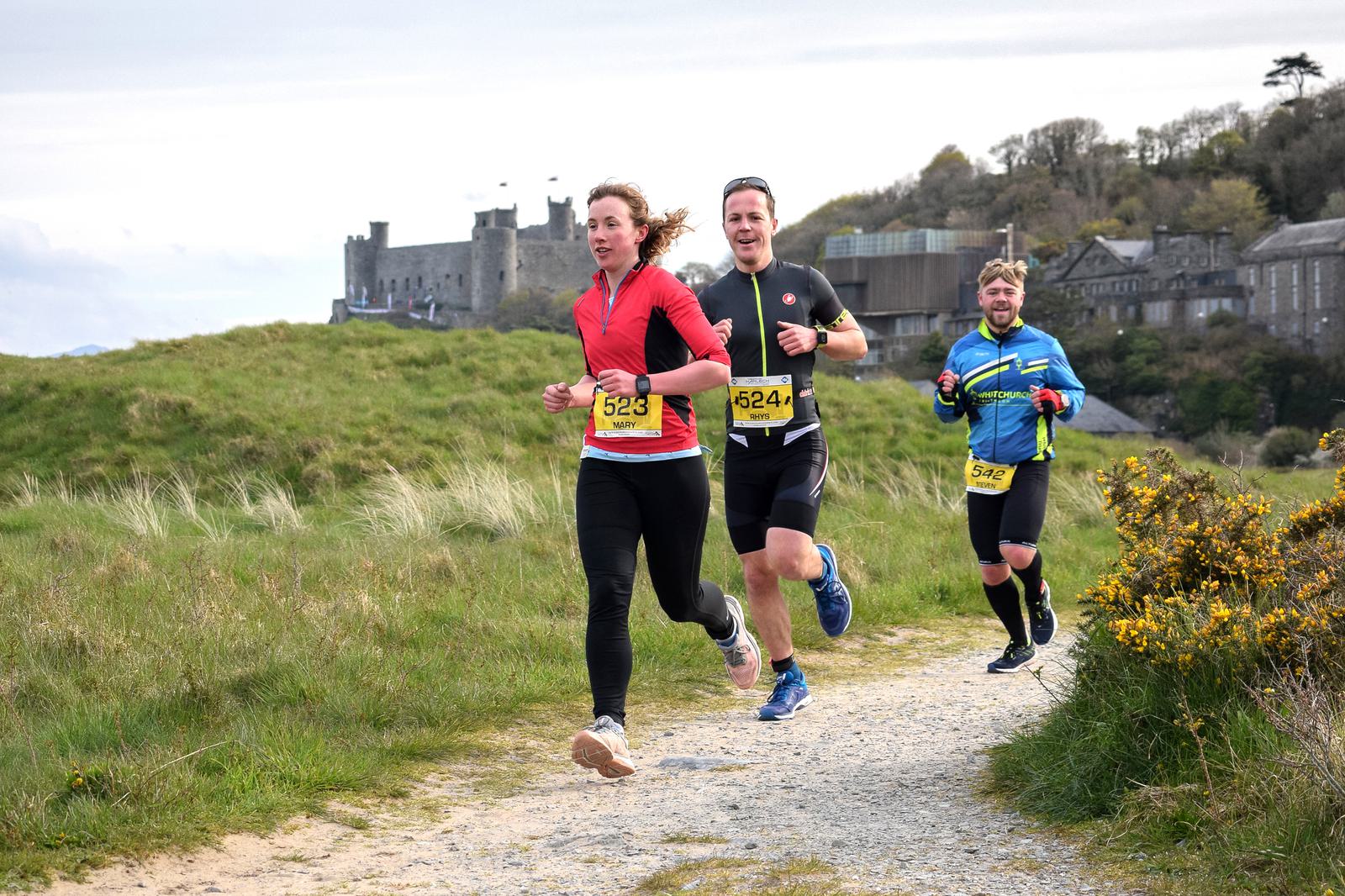 Three runners are jogging on a gravel path through a green field, with an old stone castle visible in the background. The first runner wears a red long-sleeve top, the second a black top, and the third a blue jacket. All have yellow numbered race bibs.