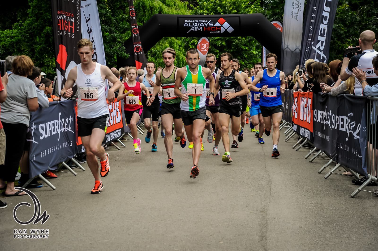 A group of runners participating in a race, wearing numbered bibs, running past banners and barriers. Spectators stand on either side of the course, cheering them on. Trees and an archway with race logos are visible in the background.