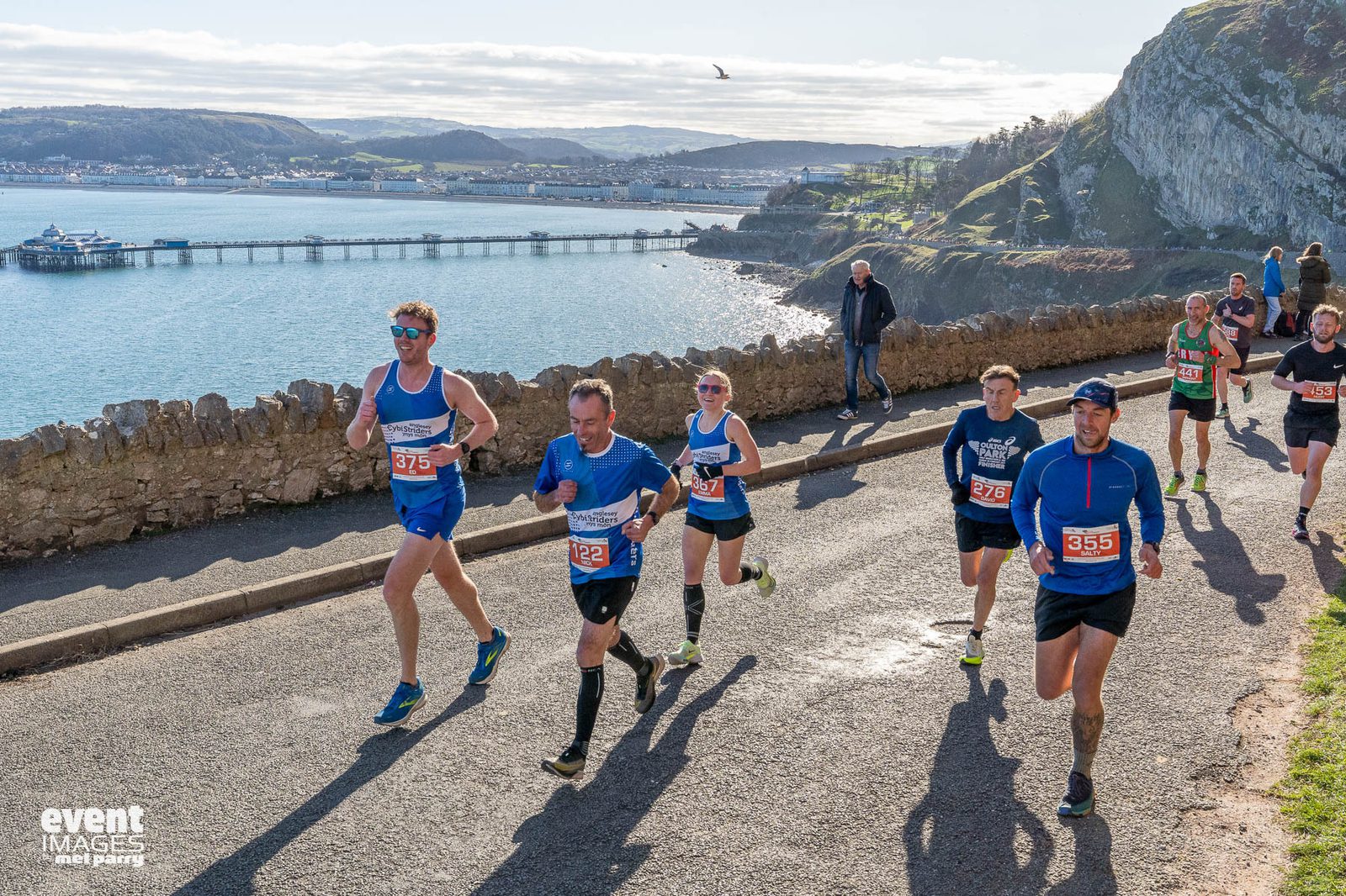 Runners participate in a road race along a scenic coastal route, with a stone wall, water, and hills in the background. The sun is shining, and race bibs are visible on the runners’ shirts.