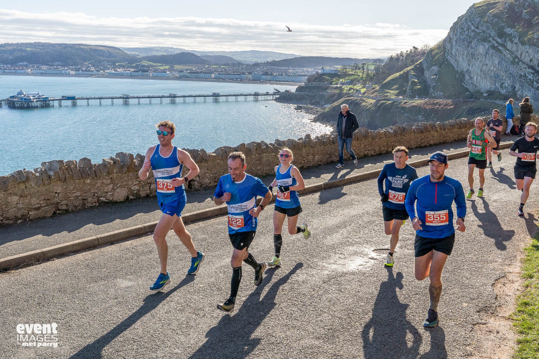 Runners participate in a road race along a scenic coastal route, with a stone wall, water, and hills in the background. The sun is shining, and race bibs are visible on the runners’ shirts.