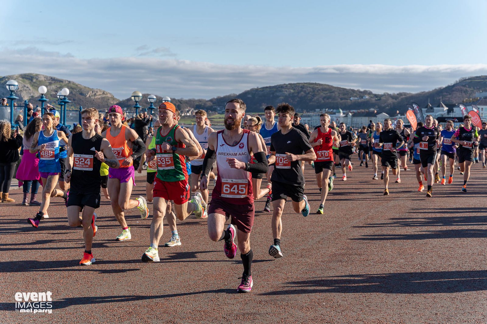 A large group of runners in numbered bibs race outdoors on a wide path with scenic hills and buildings in the background. The weather is clear and sunny. Spectators and event flags are visible along the sides.