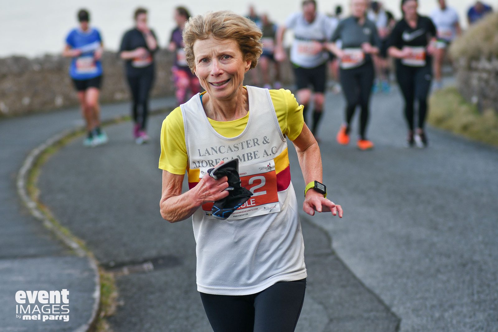 A woman in a race bib runs uphill during a road race, smiling and holding a small item in her hand. Other runners are visible in the background. The event logo and photographer credit are in the corner.