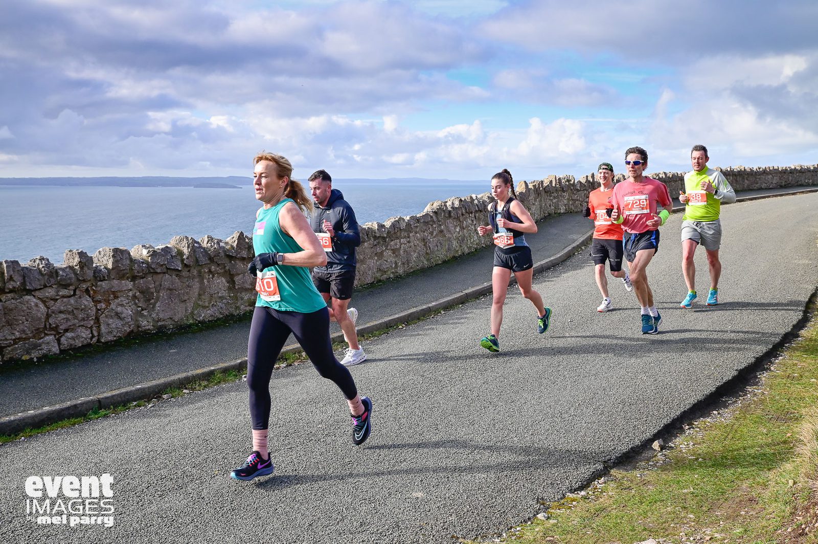 Six runners participate in a road race along a coastal path with stone walls, ocean, and clouds visible in the background. Most wear race bibs and athletic gear, running uphill on a sunny day.