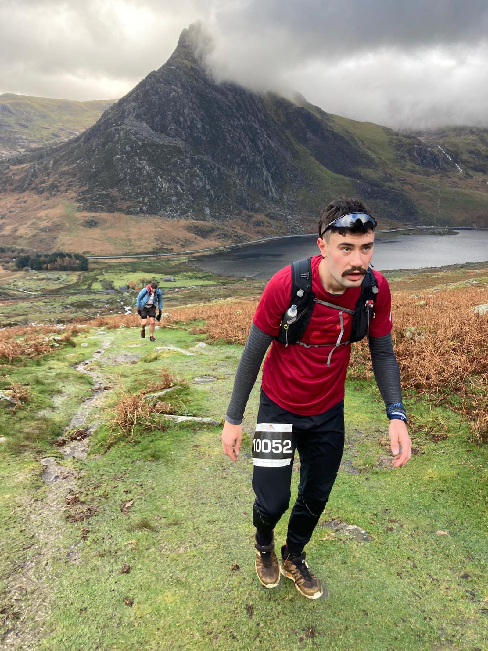 A runner wearing a red shirt, dark pants, and a race bib numbered 10052 treks up a rugged trail with a mountainous backdrop. Another runner is visible in the distance. The scene is set in a mountainous area with sparse vegetation and a lake in the background.