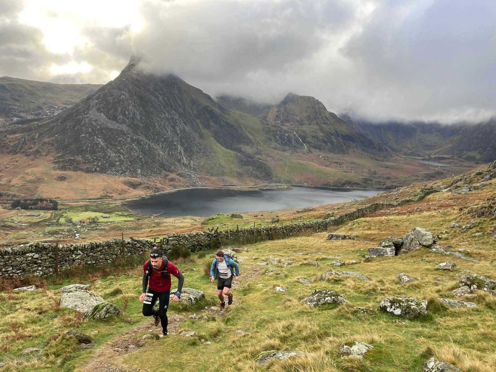 Two runners are climbing a grassy, rocky hill with a serene lake and towering mountains in the background. The sky is overcast with clouds partially obscuring the peaks. One runner is wearing a red jacket and black shorts, and the other has a blue shirt and black shorts.