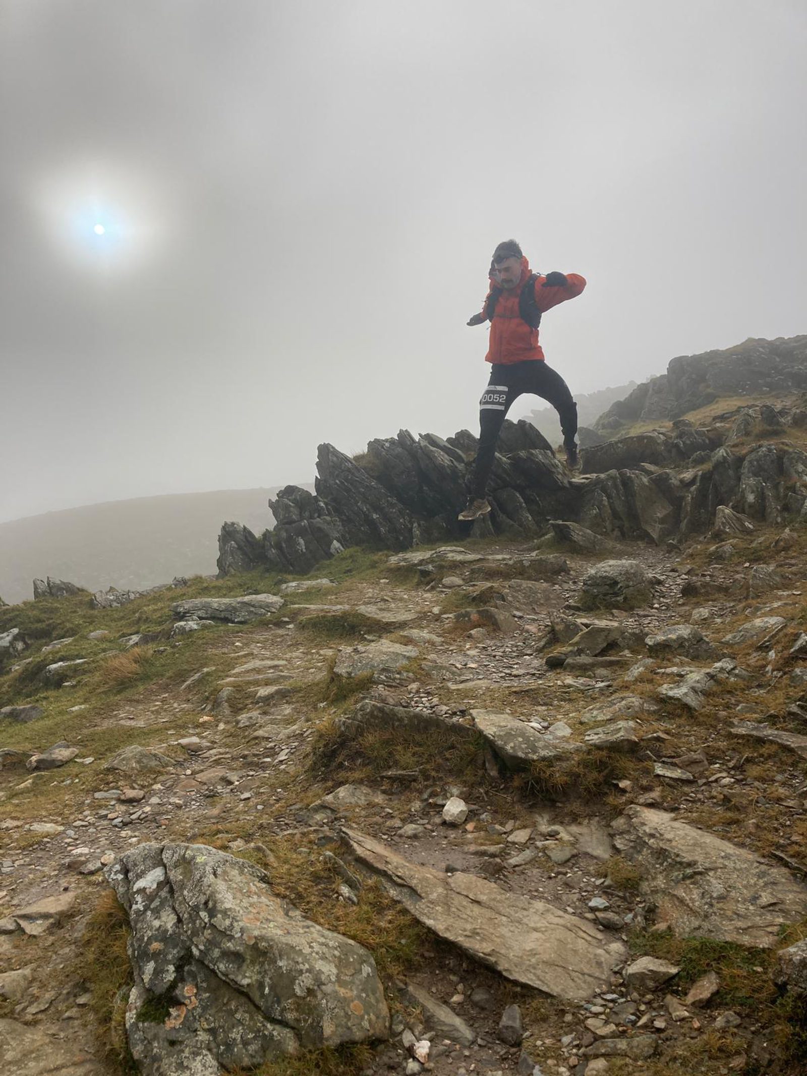 A person wearing a red jacket and dark pants is hiking on a rocky mountain trail on a foggy day. The hiker is captured mid-step, with a slight leap, and appears to be navigating through jagged rocks. The sky is overcast, and the terrain is rugged.
