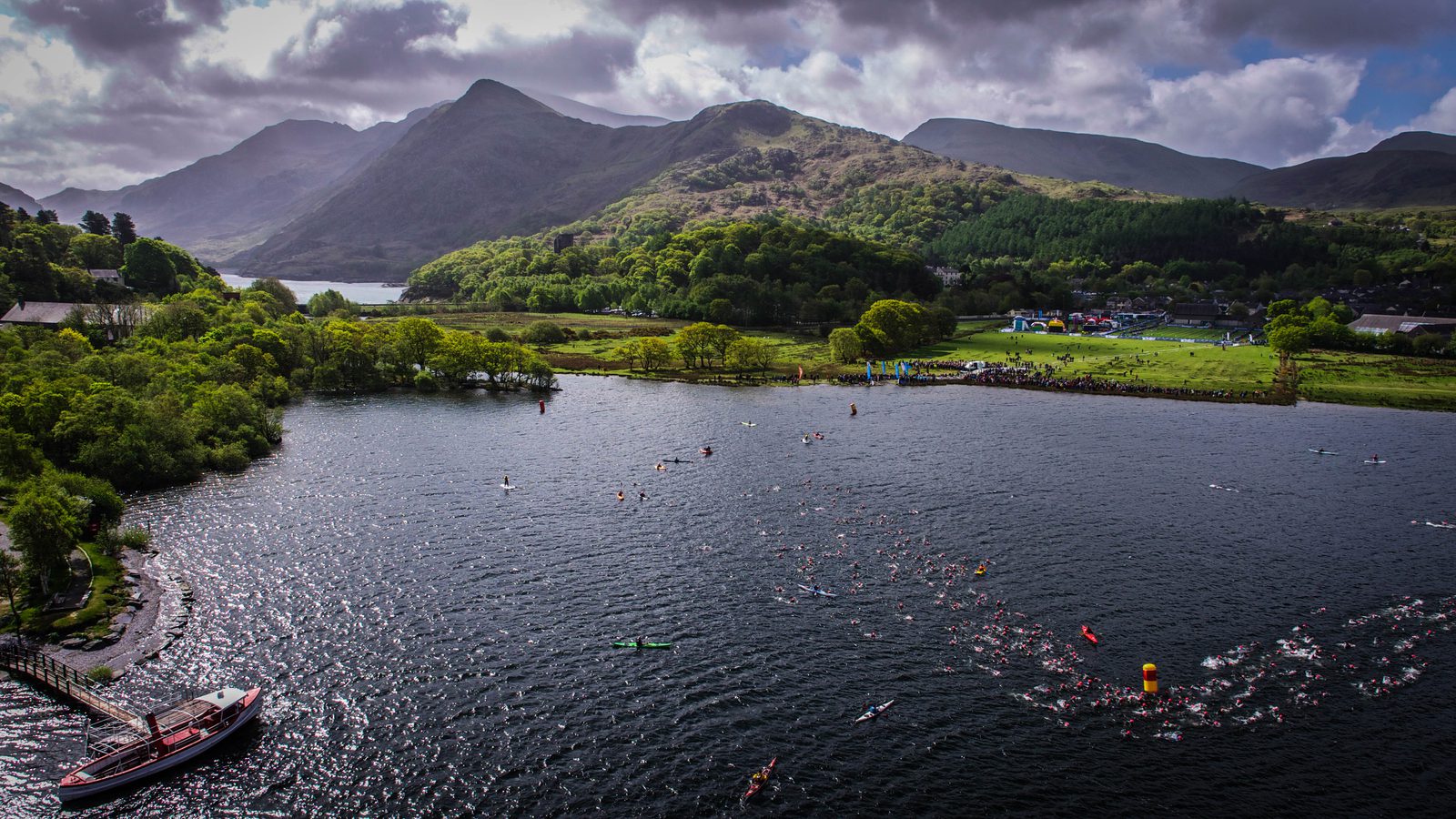 Aerial view of a large lake surrounded by lush, green hills and mountains under a cloudy sky. Numerous swimmers participate in an open-water event, with a few kayaks and a boat nearby. Onshore, tents, trees, and buildings are visible.