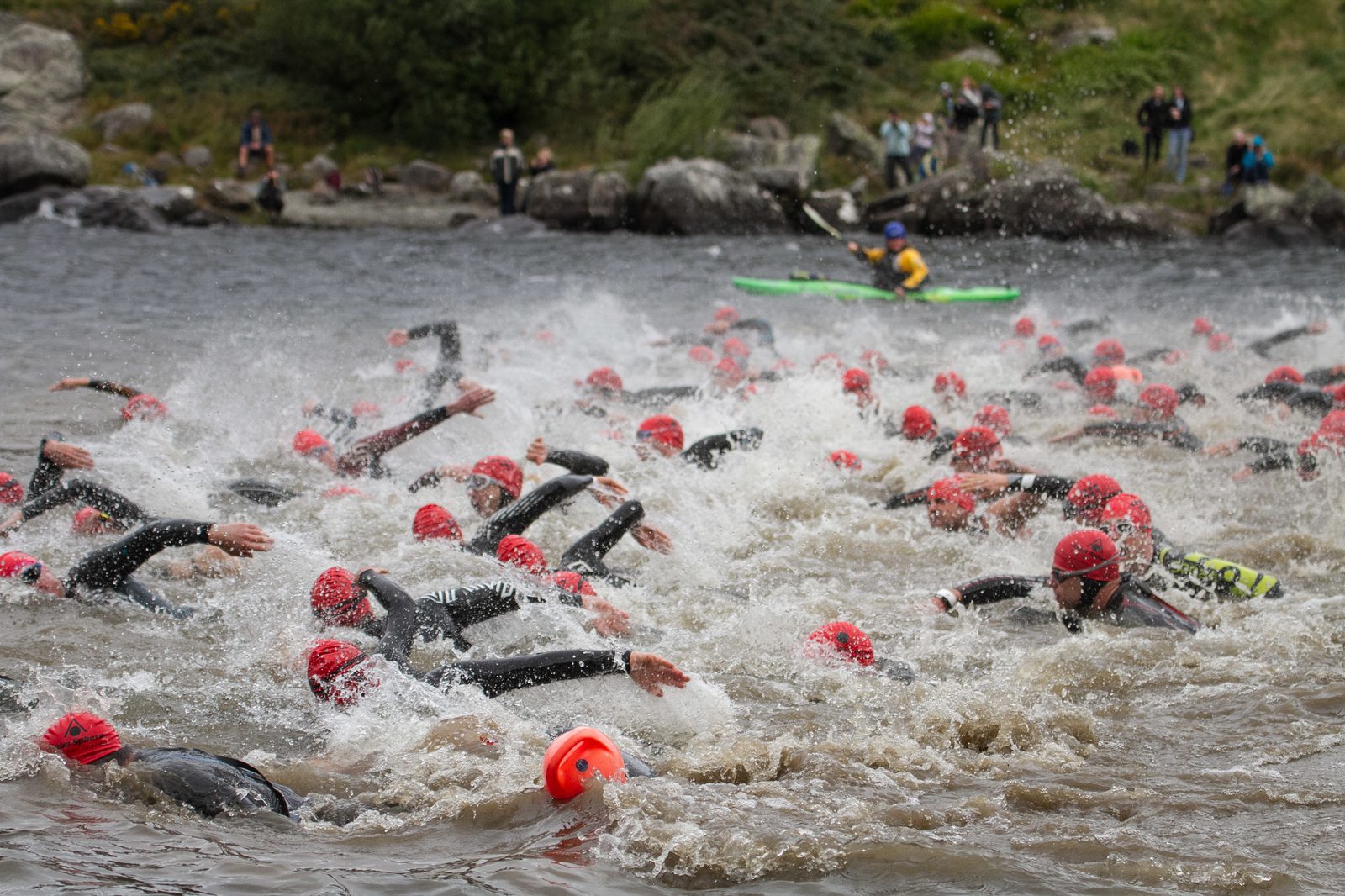 A large group of swimmers wearing red caps and wetsuits participates in an open-water race. They create splashes as they swim through choppy water. In the background, spectators watch from the shore, and a kayaker in a yellow vest helps oversee the event.